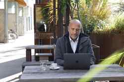 Ein Mann sitzt auf einem Holztisch eines Cafes und schaut in seinen vor sich geöffneten Laptop.
