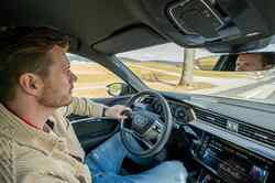 Ein Mann sitzt auf dem Fahrersitz eines Audis und hält das Lenkrad mit einer Hand während er aus der Windschutzscheibe schaut.