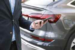 Ein Mann in Anzug hält sein Smartphone in der Hand. Hinter ihm ist ein graues Auto zu sehen.