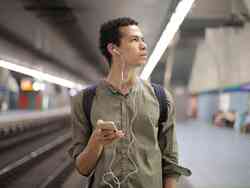 Ein Mann mit Kopfhörern und seinem Smartphone in der Hand steht an einer U-Bahn Station und schaut fragend nach rechts.