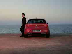 Ein Mann lehnt außen an der Fahrertür eines roten SEAT Leon alleine an einem Strand in der Abenddämmerung
