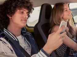 Mann und Frau sitzen gemeinsam im Auto. Während die Frau fährt schaut der Mann auch sein Smartphone.