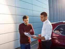 Zwei Männer stehen vor einem roten Auto und schauen sich Dokumente an
