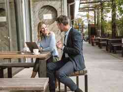 Ein Mann und eine Frau sitzen an einem Tisch in einem Restaurant und unterhalten sich. Vor Ihr steht ein aufgeklappter Laptop.
