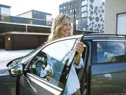 Eine Frau steht in der offenen Fahrertür eines grauen Autos und lächelt.