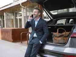 Ein Mann lehnt im Kofferraum von einem grauen Auto und telefoniert.