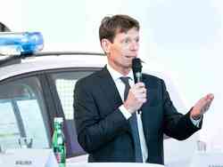 Hannes Maurer, CEO der Porsche Bank AG, steht vor einem Polizeiwagen mit einem Mikrofon in der Hand und hält eine Rede.