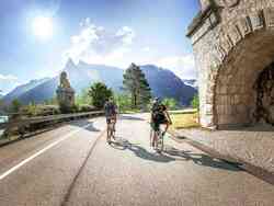 Zwei Männer fahren mit dem Fahrrad einen Weg hinauf. Vor ihnen sind Berge zu sehen.