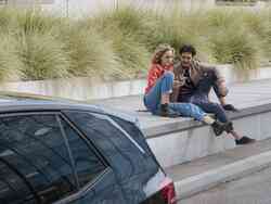 Junges Pärchen aus Mann und Frau sitzen auf Stufen hinter einem dunkelblauen Auto