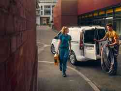 Ein Mann steht neben der offenen Tür des Kofferraums eines weißen VW Caddy Cargo. Eine blonde Frau geht an ihm vorbei und schaut in seine Richtung.