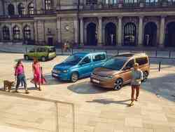 Ein blauer und ein bronzefarbener VW Caddy parken, zwei frauen sowie ein Mann spazieren an dem Fahrzeug vorbei
