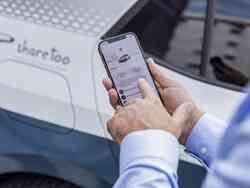 Ein Mann tippt auf seinem Smartphone. Hinter ihm steht ein weißes Fahrzeug mit der Aufschrift "sharetoo"