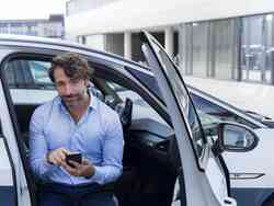 Ein Mann sitzt auf dem Beifahrersitz eines weißen Autos und schaut auf sein Smartphone. Die Beifahrertür ist offen.