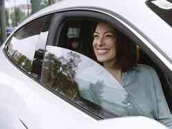 Eine Frau sitzt auf dem Beifahrersitz eines weißen Autos und lächelt aus dem halb geöffneten Fenster.