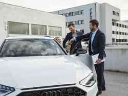 Zwei Männer in Anzug stehen neben einem weißen Audi, in welchen eine blonde Frau gerade einsteigt.