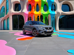 Seitliche Frontansicht des SEAT Arona, der vor einem Gebäude parkt. Das Gebäude und der Boden sind von bunten, verlaufenden Farben überzogen